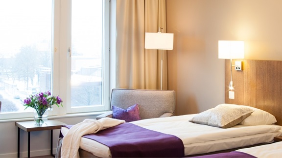 Bilden föreställer ett hotellrum på Plaza. I förgrunden en säng bäddad med lila överkast och ett bord med en vas med lila blommor.