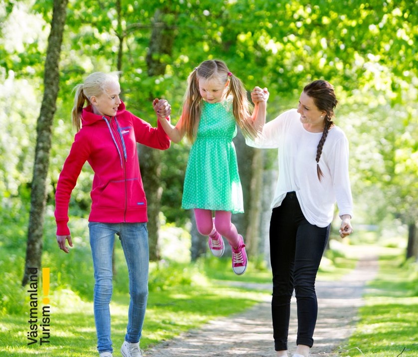 Bilden föreställer en kvinna och två barn som promenerar på en stig i ett grönområde. 